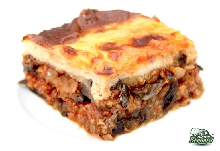 Une vue rapprochée d'une tranche de moussaka, mettant en valeur la cuisine grecque traditionnelle avec sa composition en couches de pommes de terre, d'aubergines, de viande hachée et de sauce béchamel crémeuse sur le dessus, sur une assiette blanche.