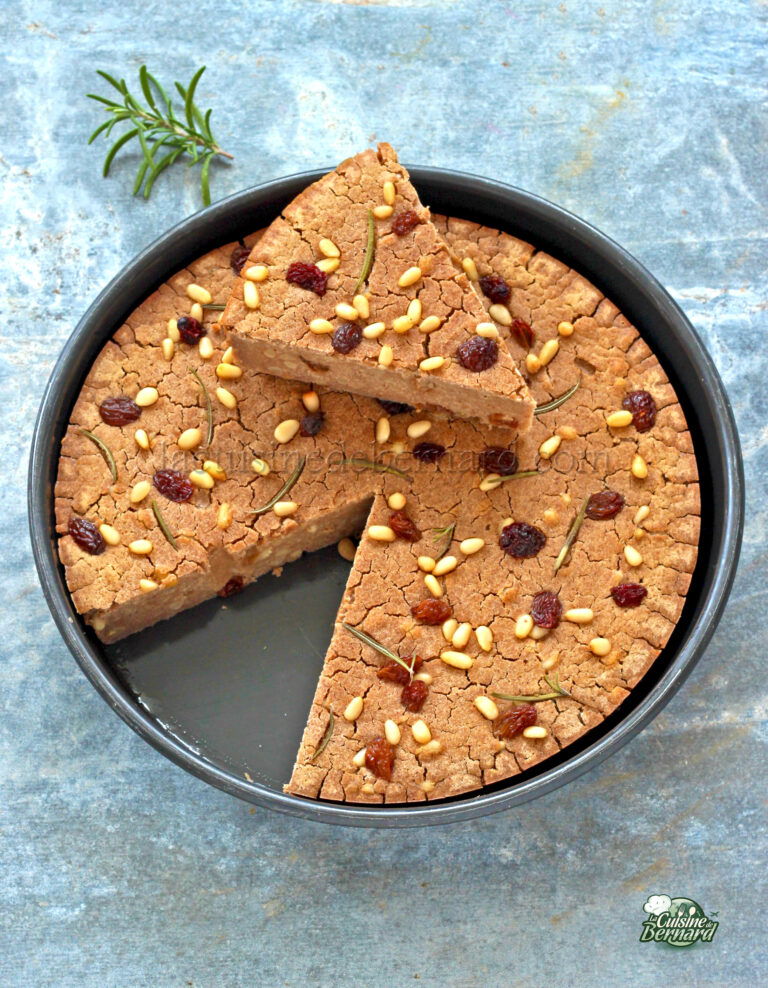 A round cake with nuts and dried fruits on top, sliced into wedges on a blue textured background.