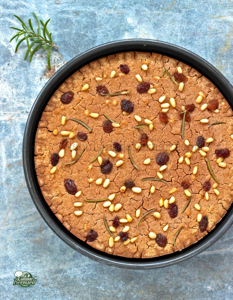 A freshly baked round cake with pine nuts and rosemary on top, served on a blue textured surface.