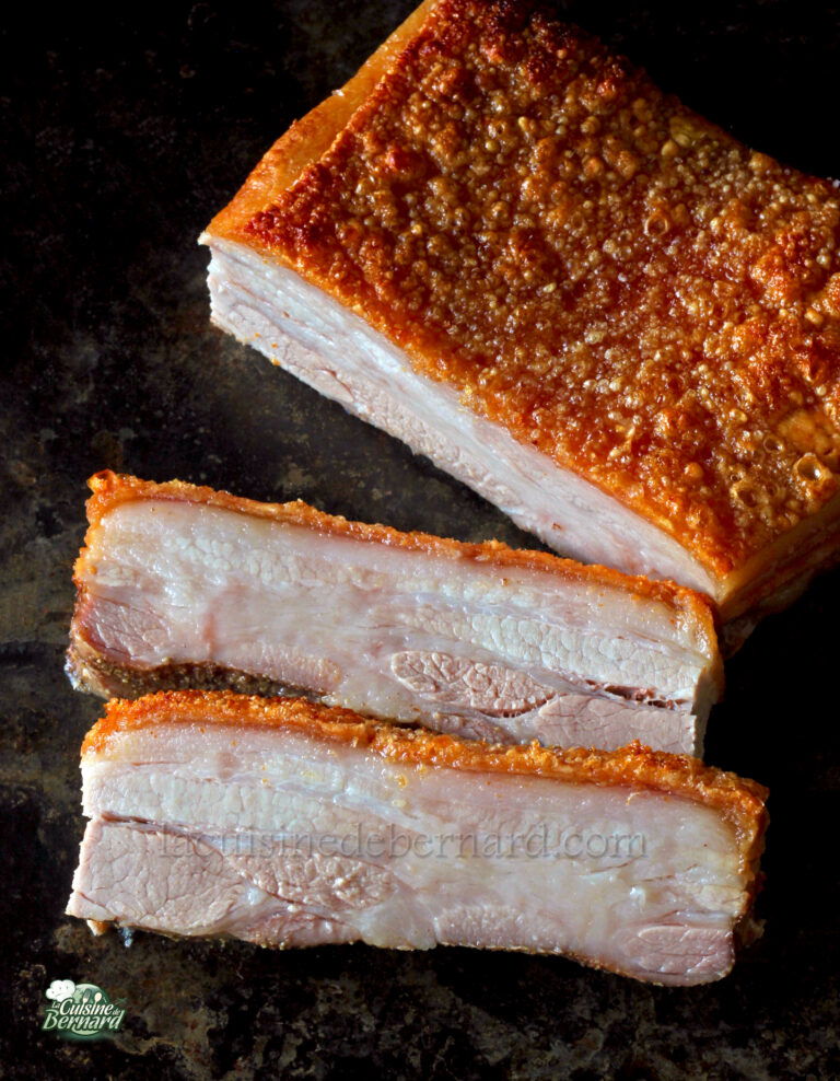 A slab of cooked pork belly with crispy skin sits on a dark surface. Two slices are cut from the slab, revealing the juicy meat inside.