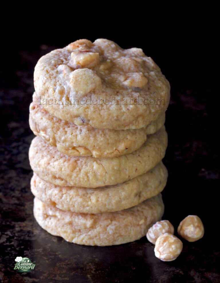 A stack of five cookies with nuts on a dark surface, with two nuts placed beside the stack. The cookies have a homemade appearance.