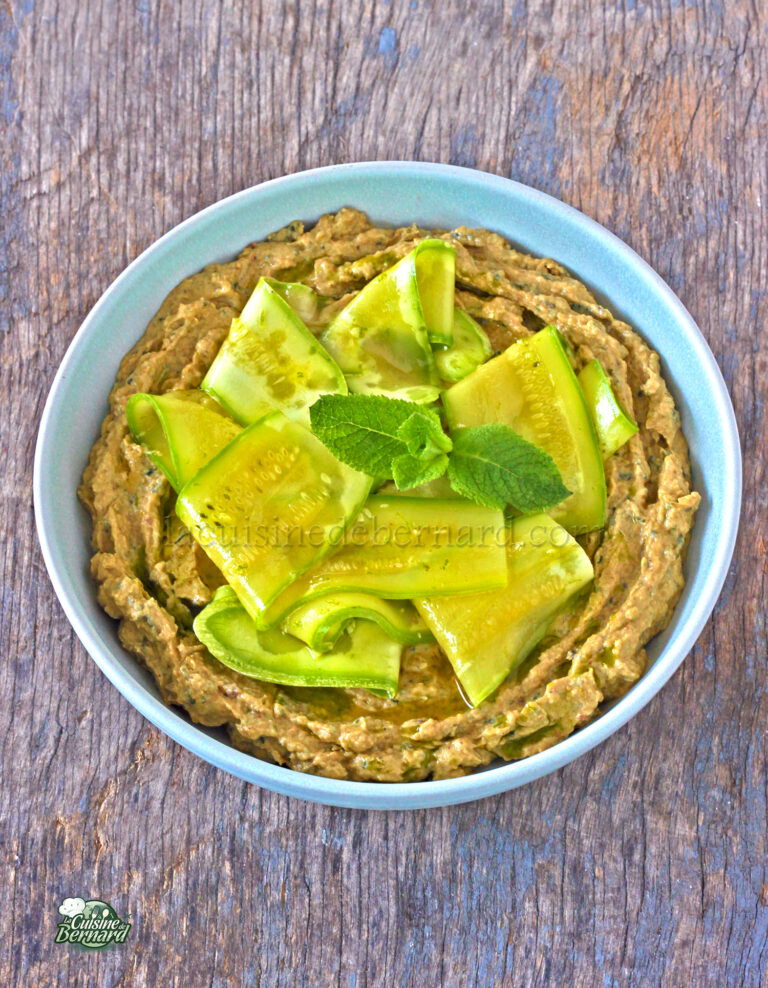 A bowl of hummus garnished with cucumber slices and a sprig of mint, served on a wooden surface.