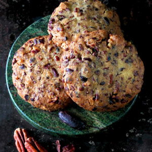Trois gros biscuits aux noix de pécan empilés sur une assiette texturée vert foncé, entourés de pacanes et de canneberges en vrac, sur un fond sombre.