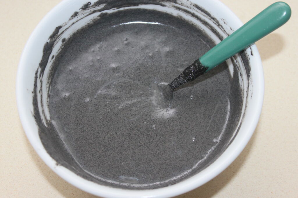 Bol blanc contenant un mélange liquide gris, rappelant le tahiné noir, avec une cuillère à manche vert partiellement enduite du mélange.