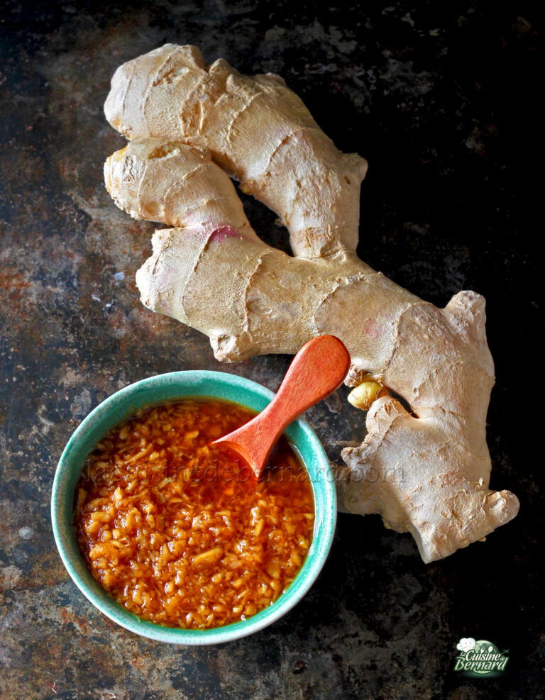 A piece of whole ginger root lies next to a small bowl of finely chopped ginger mixed in a red-brown liquid, with a wooden spoon resting in the bowl.