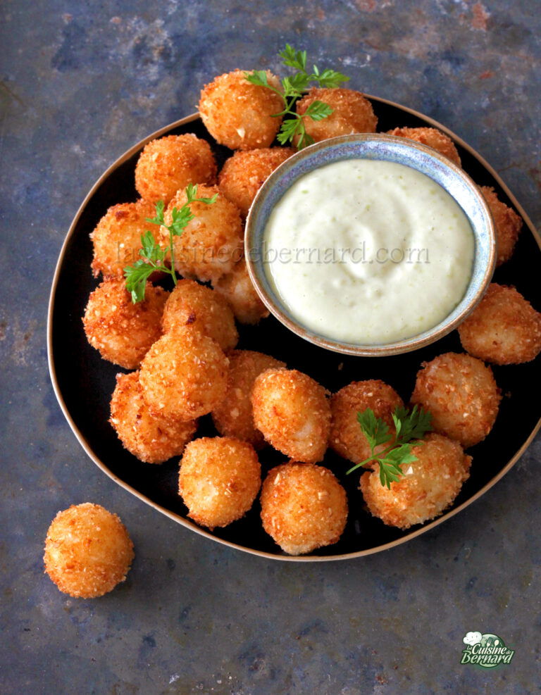 A plate of breaded and fried appetizer balls garnished with parsley, accompanied by a small bowl of dipping sauce. The plate is on a dark, textured surface.