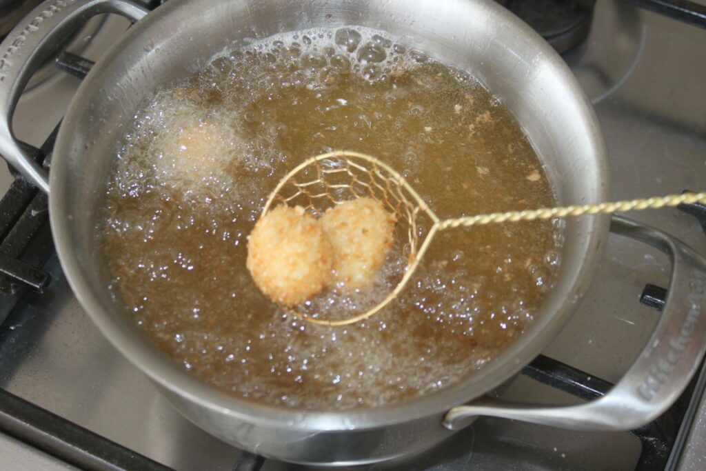 Uma escumadeira de metal retira duas bolas de mozza empanada do óleo quente em uma frigideira no fogão.