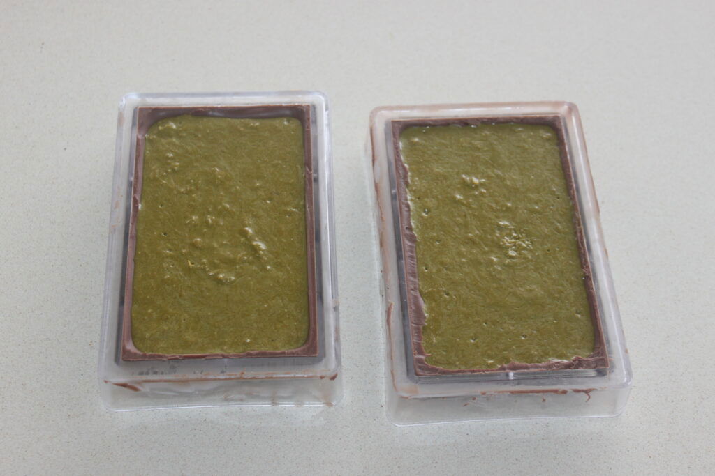 Deux moules rectangulaires remplis d'un mélange vert, partiellement entourés d'une couche de chocolat de luxe, sont placés sur un plan de travail de couleur claire.