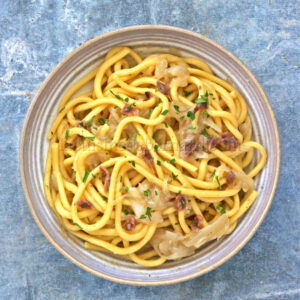 Un bol de pâtes Bigoli aux nouilles épaisses, garni d'herbes et mélangé à des oignons caramélisés et des petits morceaux de viande. Le plat est joliment présenté sur une surface grise texturée, subtilement rehaussée d'une touche de salsa pour une saveur plus intense.