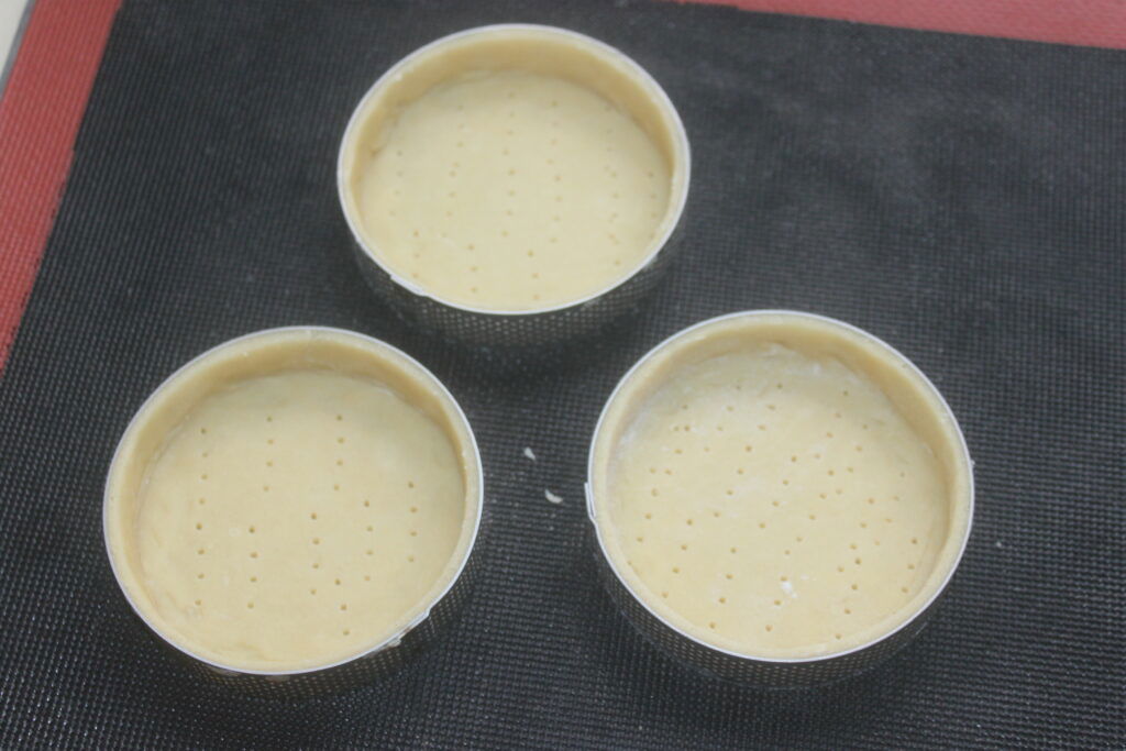 Trois tartelettes crème d'orange crues aux fonds piqués sont disposées sur un plan noir, prêtes à être garnies.