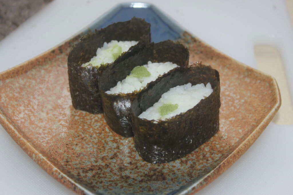 Trois morceaux de sushi enveloppés d'algues, remplis de riz et garnis d'une petite quantité de wasabi, servis sur une assiette en céramique brune.