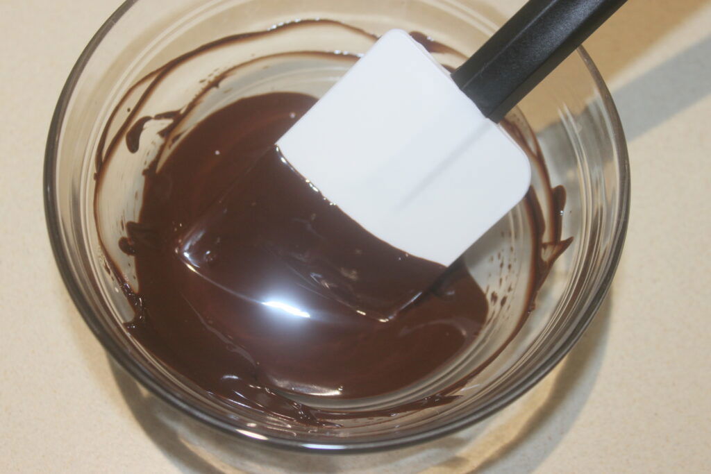 Un bol en verre contenant du chocolat fondu avec une spatule posée sur le bord, parfait pour arroser les sablés ou les amandes grillées.