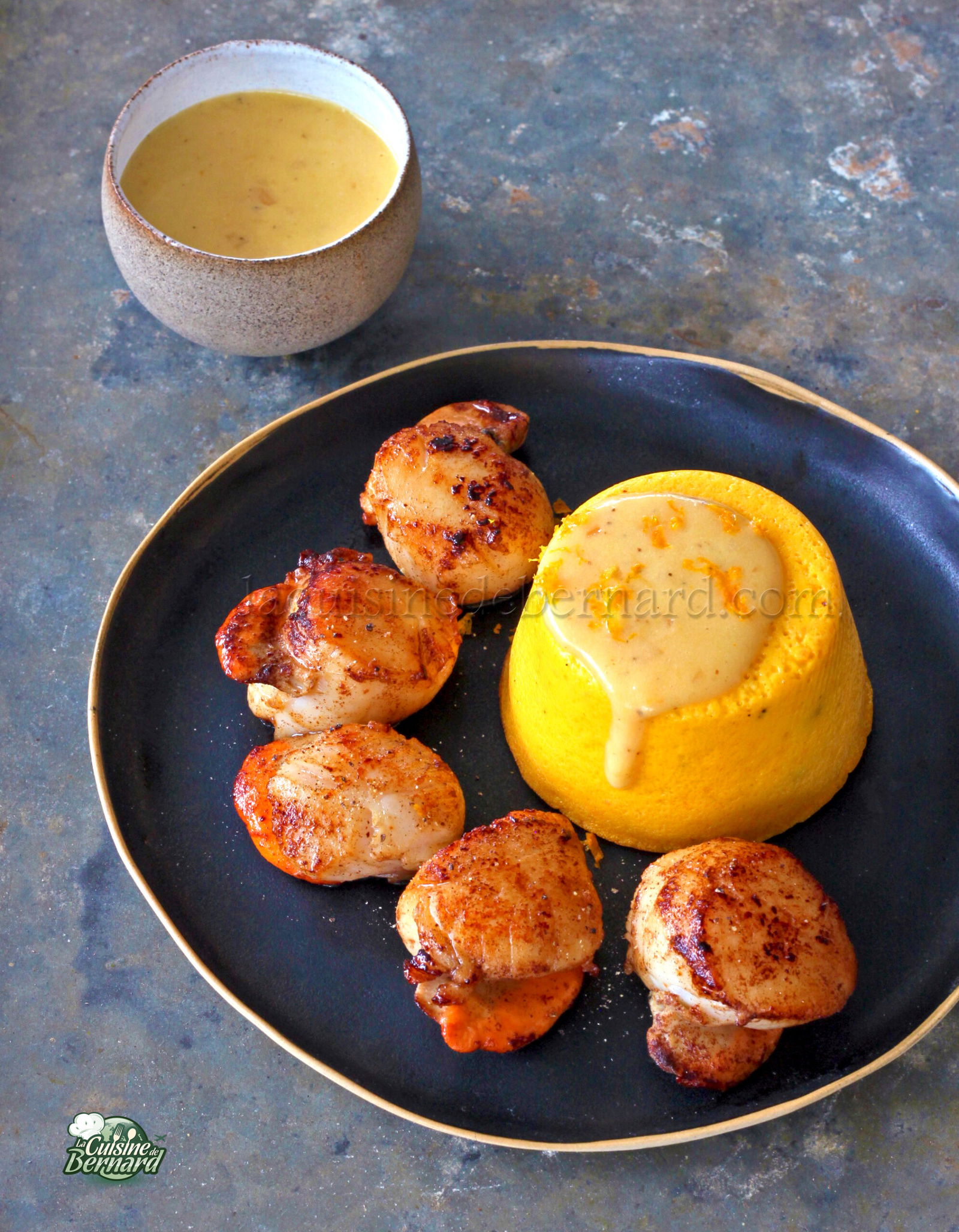 Des coquilles Saint Jacques grillées élégamment présentées sur une assiette noire, accompagnées de beurre à l'orange et d'un flan de potimarron vibrant. Une tasse de sauce supplémentaire trône en arrière plan, rehaussant l'attrait du plat.