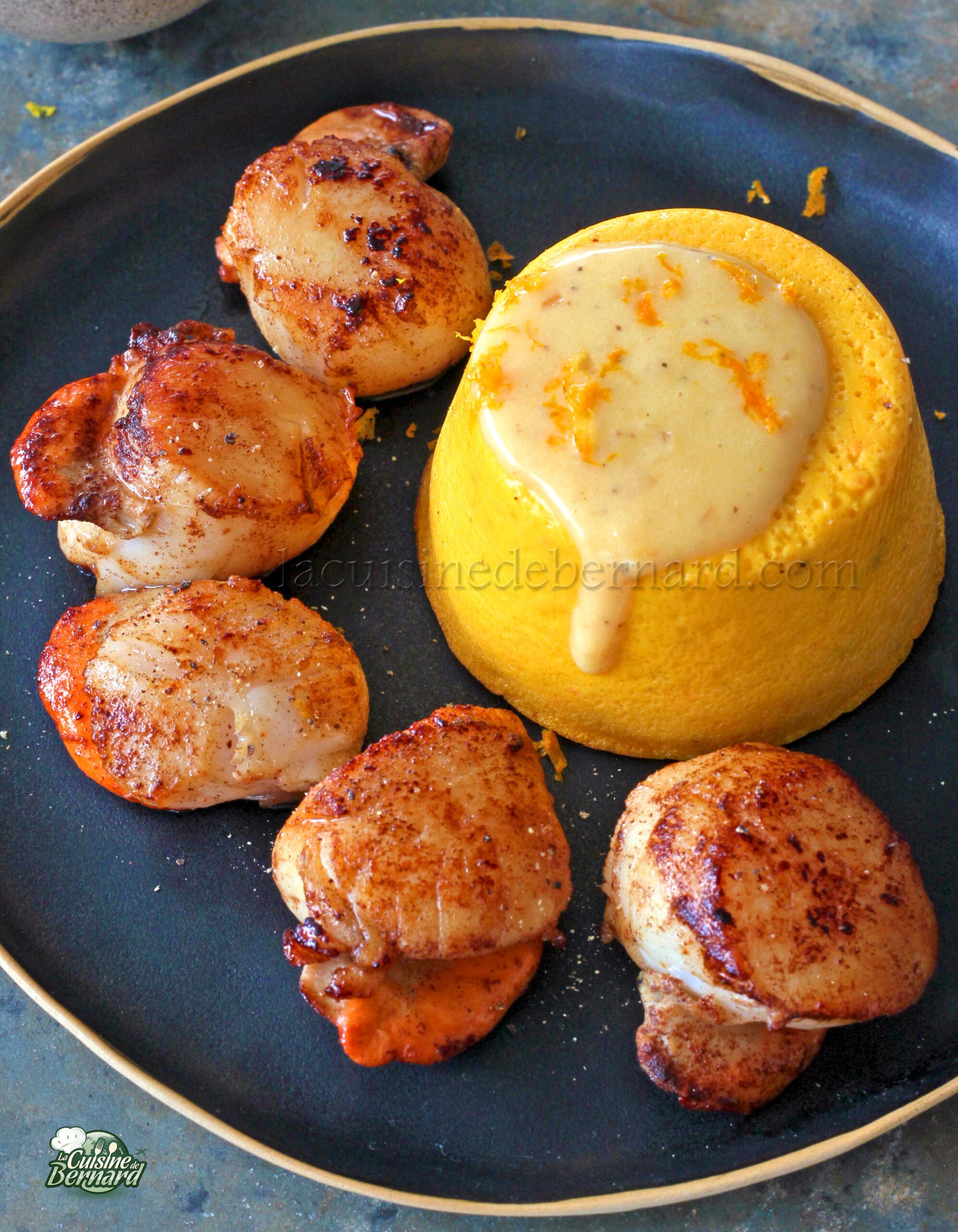 Des coquilles Saint Jacques poêlées reposent sur une assiette noire à côté d'un flan de potimarron, nappé d'une sauce crémeuse au beurre à l'orange.