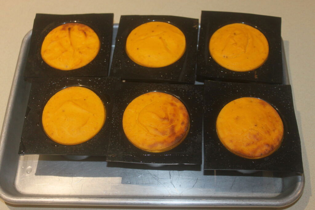 Six muffins ronds au potimarron orange, emballés dans des emballages noirs, sont élégamment disposés sur un plateau en argent, offrant une délicieuse touche de beurre à l'orange.