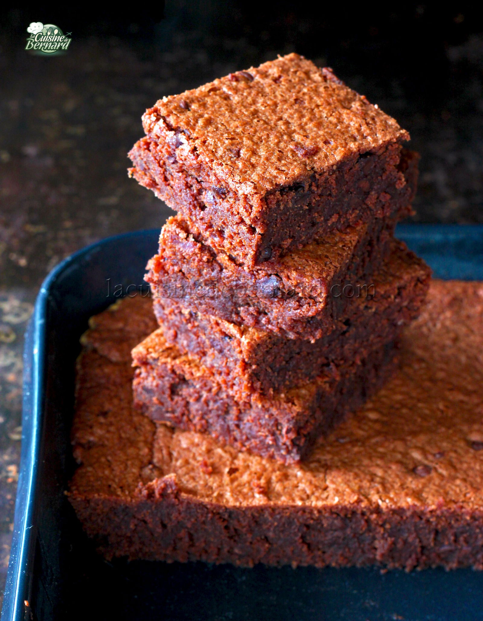 Une pile de quatre brownies au chocolat avec une surface texturée et friable se trouve sur la plaque de cuisson, rappelant un gâteau au chocolat décadent.