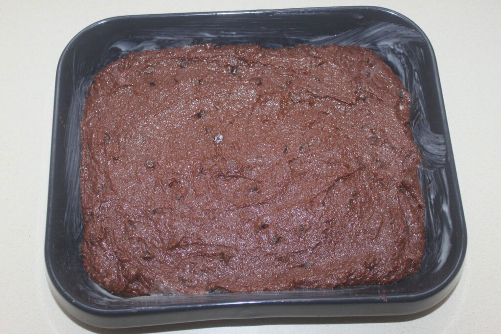 Un moule carré rempli de pâte à brownie au chocolat non cuite saupoudrée d'amandes croquantes repose sur un plan de travail clair.