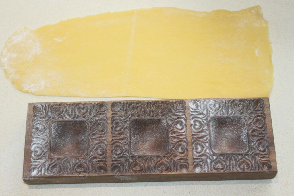 Un moule à raviolis en bois aux motifs décoratifs repose à côté d'une feuille de pâte à pâtes roulée, prêt à confectionner de délicieux raviolis à la ricotta sur une surface claire.