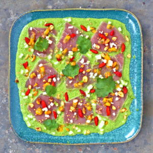 Une assiette carrée bleue présente des tranches de thon cru sur une sauce en purée verte, garnie de piments rouges, de noix et de feuilles de coriandre, évoquant une touche fusion au ceviche traditionnel.