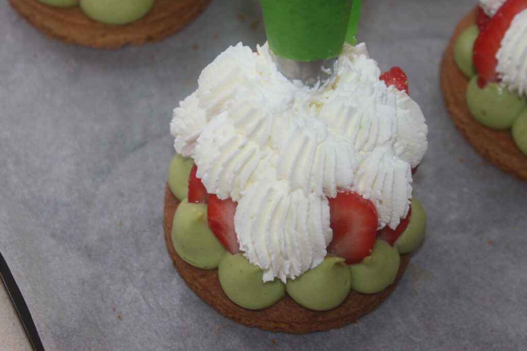 Une pâtisserie à la crème basilic verte et aux fraises fraîches est élégamment garnie de chantilly mascarpone à l'aide d'une poche à douille. Les délicieuses tartelettes fraise reposent sur une plaque à pâtisserie tapissée de papier sulfurisé.