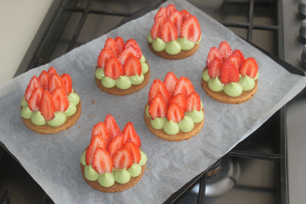 Six biscuits garnis de crème de matcha vert et de tranches de fraises sont posés sur une plaque à pâtisserie tapissée de papier sulfurisé sur une cuisinière, ressemblant à de délicieuses tartelettes fraîches.
