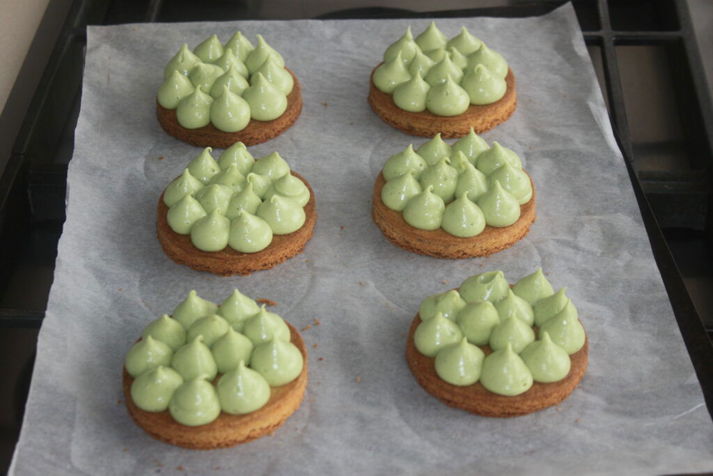 Six biscuits ronds avec des cuillerées de crème verte sur papier sulfurisé, disposés en deux rangées de trois, sur une plaque à pâtisserie, rappelant d'élégantes tartelettes fraîches avec des touches de chantilly mascarpone et de crème basilic chocolat blanc.