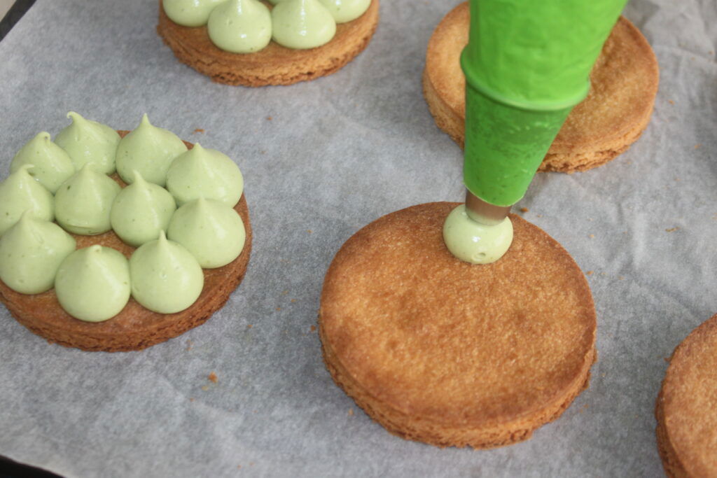 Gros plan d'un glaçage vert déposé sur des biscuits ronds cuits sur du papier parchemin. D'autres biscuits au glaçage vert passepoilé sont visibles à proximité, ressemblant aux détails complexes de la crème basilic chocolat blanc.