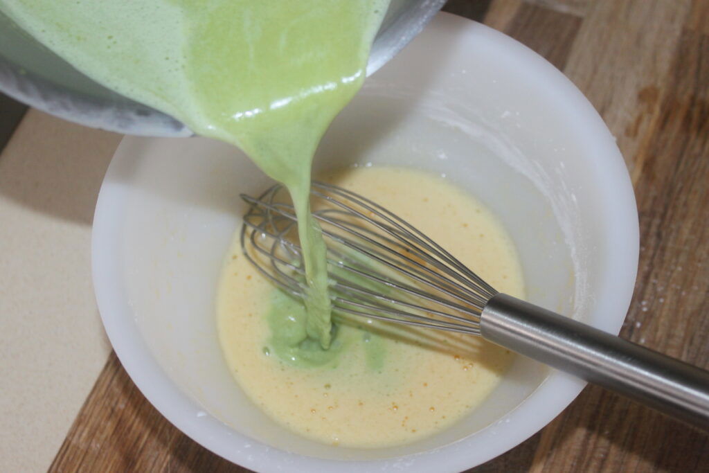 Le liquide vert est versé d'un récipient dans un bol blanc avec un mélange jaune et un fouet en métal à l'intérieur, parfait pour préparer la crème basilic chocolat blanc.